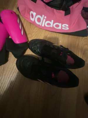 Fotbolls skor och mer - Säljer ett par svarta adidas fotbollsskor med klassiska vita ränder på sidan och rosa detaljer på insidan. Skorna har snörning och rund tå, tillverkade i syntetmaterial som tål tuffa matcher. Perfekta för dig som älskar fotboll och vill ha stil på planen.