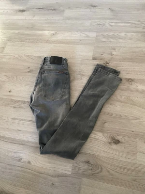 Grå  från Nudie Jeans - Snygga grå slim fit  jeans från Nudie Jeans med klassisk femficksdesign och svarta detaljer på bakfickan. Jeansen har en smal passform och är tillverkade i mjukt jeanstyg som sitter tajt längs benen. Perfekt för dig som gillar en stilren och modern look.