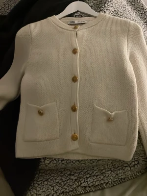 Vit stickad kofta från Zara S - Säljer en vit stickad kofta från Zara i storlek S. Koftan har guldfärgade dekorativa knappar framtill och på fickorna, rund halsringning och två fickor fram. Perfekt till lager på lager och ger en clean, klassisk look.