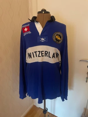 Blå piké Polo Ralph Lauren Switzerland - Blå långärmad pikétröja från Polo Ralph Lauren med svart krage, vit bred rand över bröstet och texten SWITZERLAND. Broderade märken och detaljer framtill, stor romersk siffra IV på ryggen. Skön bomull, perfekt för dig som gillar sportig stil. Säljes på grund av att den har blivit för liten!