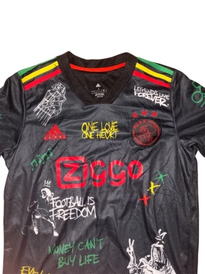 Ajax x Bob Marley Adidas tröja - Unik svart Ajax x Bob Marley tröja från Adidas med färgglada detaljer i rött, gult och grönt på axlar och ärmslut. Tryck med Bob Marley, Ajax-logga, Ziggo och flera inspirerande texter och illustrationer både fram och bak. Perfekt för fotbollsfans och streetwear.
