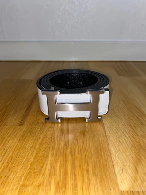 Svart och vit skinnbälte med H-spänne - Snyggt bälte i svart och vitt skinn med ett stort metallspänne i form av ett H. Bältet har en stilren design med tydliga sömmar och struktur i lädret. Perfekt för att lyfta din outfit med en cool detalj.