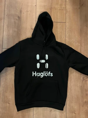 Haglöfs hoodie - Svart (helt ny) - Säljer en helt ny och oanvänd hoodie från Haglöfs i klassisk svart färg. Stilren design med stort Haglöfs-logo tryck framtill. Hoodien har huva med dragsnören samt känguruficka framtill.  Tillverkad i ett mjukt och tjockt material som ger hög komfort och värme – perfekt för vardag, träning eller chill. Tidlös modell som aldrig går ur stil.  Skick: Helt ny, aldrig använd Färg: Svart Detaljer: Huva, känguruficka, tryck framtill       Passform: Bekväm / normal