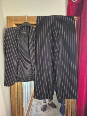 Svart randig kostym från Rut&Circle - Säljer en svart kostym från Rut&Circle i storlek 36. Setet består av en kavaj och ett par raka byxor, båda med vita smala ränder. Materialet är mjukt och har en lätt glansig finish på insidan av kavajen. Perfekt för dig som gillar stilrena och trendiga looks. Byxorna är från gina tricot