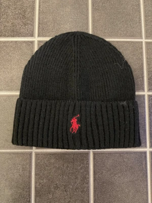 Svart stickad mössa från Polo Ralph Lauren - Svart ribbstickad mössa från Polo Ralph Lauren med röd broderad logga framtill. Klassisk och enkel design som passar perfekt till kyliga dagar.