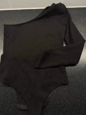 Svart oneshoulder body från Lost Ink - Säljer en svart body från Lost Ink med oneshoulder-design och lång ärm. Kroppsnära passform och knäppning nedtill. Materialet är mjukt och stretchigt med 63% viskos, 32% nylon och 5% elastan. Perfekt till svarta jeans eller kjol för en cool look.