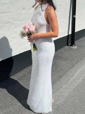 Vit långklänning med öppen rygg - Säljer en elegant vit långklänning med halterneck och öppen rygg. Klänningen har en slits bak och är figurnära med ett skimrande tyg som ger en lyxig känsla. Perfekt för speciella tillfällen och sommarens höjdpunkter. Inga skavanker 