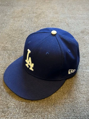 Mörkblå LA 59FIFTY keps från New Era - Säljer en mörkblå LA-keps från New Era, modell 59FIFTY. Klassisk rak skärm och broderad LA-logga framtill i vitt. Tillverkad i 100% polyester och har MLB-logga bak. Perfekt för dig som gillar streetwear och sportig stil.