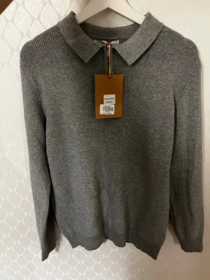 Grå stickad pikétröja med half zip - Säljer en helt ny oanvänd stilren grå stickad pikétröja från Cattaleya i storlek S-M. Tröjan har klassisk krage, quarter zip-dragkedja framtill och ribbade muddar vid ärmslut och nederkant. Perfekt för dig som gillar en clean och enkel look.