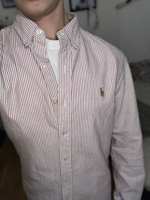 Randig skjorta från Polo Ralph Lauren - Säljer en klassisk skjorta från Polo Ralph Lauren med smala röda och vita ränder. Skjortan har lång ärm, knappar hela vägen och ikonisk broderad logga på bröstet. Tillverkad i mjuk bomull och har en normal passform. Perfekt för dig som gillar stilrena och tidlösa plagg. Storlek S, jag är 177 cm och väger 68 kg och den passar utmärkt. Såklart äkta. Inga som helst defekter, knappt använd. Nypris 1300. Mitt pris 500. Bara att höra av er!