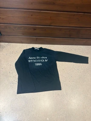 Svart långärmad topp Acne Studios - Säljer en svart långärmad topp från Acne Studios med tryck framtill där det står 'Acne Studios STOCKHOLM 1996'. Toppen har en enkel, rak passform och är gjord i mjuk bomull. Perfekt till en clean och stilren look.