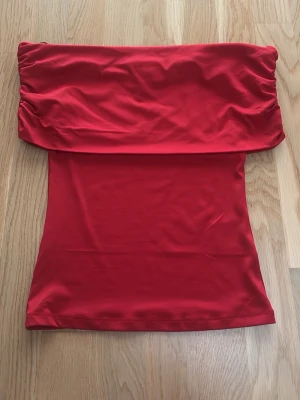 Röd bandeau tubtopp - Säljer en röd bandeau tubtopp med snygga rynkade detaljer upptill. Toppen är axelbandslös och har en slät, tight passform. Materialet känns mjukt och stretchigt, perfekt för sommarens alla tillfällen. Färgen är intensivt röd och ger en riktigt poppig look.