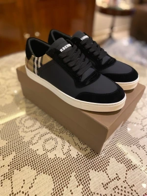 Burberry sneakers med rutig detalj - Snygga svarta sneakers från Burberry med klassisk rutig panel i beige, svart och röd på hälen. Skorna har vit platt sula, rund tå och svarta snören. Materialmix av skinn och textil, med märkets logga på tungan och bak på sulan. Perfekt för dig som gillar stilrena designer.