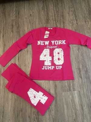 Rosa mjukisbyxor med tryck från L'Iris - Säljer ett par coola rosa mjukisbyxor från L'Iris med stort vitt tryck '48' på benet. Byxorna har en avslappnad passform och är gjorda i mjukt bomullsmaterial, perfekta för chill eller träning. Matchande långärmad tröja med 'New York Original 48 Jump Up' ingår.