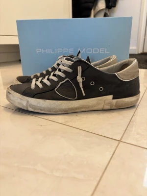 Svarta sneakers från Philippe Model - Säljer ett par svarta Philippe Model sneakers med beige detaljer och snörning. Skorna har en rund tå, platt sula och är tillverkade i skinn med inslag av mocka. Beige hälparti och vita snören ger en snygg kontrast. Perfekt för dig som gillar stilrena och coola sneakers.