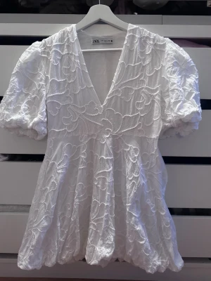 Vit kort klänning med puffärm Zara  - Säljer en vit kort klänning från Zara med v-ringning och puffiga korta ärmar. Klänningen har broderade detaljer och en lätt utsvängd kjol. Perfekt för sommarens alla tillfällen! 