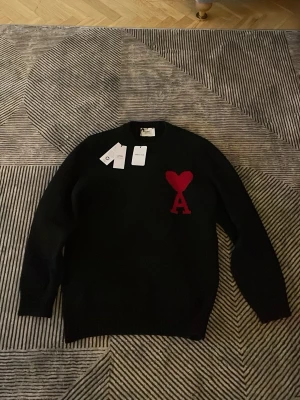 Svart stickad tröja från Ami - Svart stickad tröja från Ami med röd hjärtformad A-logga på bröstet. Tröjan har rund hals, ribbade muddar och lång ärm. Materialet är mjukt och känns lyxigt, perfekt för dig som gillar stilrena detaljer med en twist. Helt ny ami hoodie som är använd ca 1-2 gånger bara för att testa denna fina stickade tröjan kommer garanterat uppgradera din stil hör av er vid dm för frågor och funderingar!