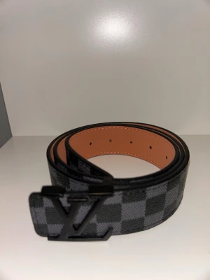 Svart och grå rutig bälte - Snyggt bälte i svart och grått med rutigt mönster och stor logga i metall framtill. Bältet är gjort i skinn med en matt svart spänne och har en lyxig känsla. Perfekt accessoar för att lyfta din outfit. Går ner i pris vid snabb affär