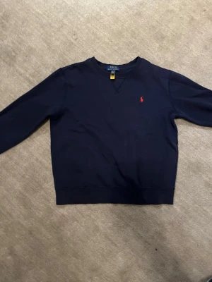 Mörkblå sweatshirt från Polo Ralph Lauren - Snygg mörkblå sweatshirt från Polo Ralph Lauren med klassisk röd broderad logga på bröstet. Tröjan har rund hals, långa ärmar och ribbade muddar vid ärmslut och nederkant. Perfekt för chill dagar och enkel att matcha med jeans eller joggers.