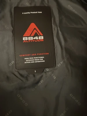 Svart och vit vindjacka från 8848 Altitude - Snygg vindjacka från 8848 Altitude i svart och vitt med röda detaljer och dragkedjor. Jackan har hög krage, flera fickor med dragkedja och coolt broderat logotyp på ärmen. Perfekt för dig som gillar äventyr och outdoor.