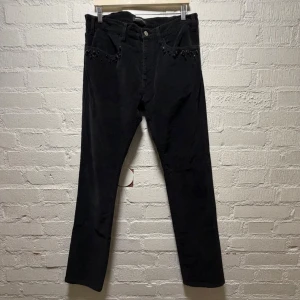 Undercover Svarta manchesterbyxor med pärlor - Svarta manchesterbyxor från Undercover i slim fit med snygga pärldetaljer vid fickorna framtill. Byxorna har klassisk femficksdesign och är tillverkade i mjuk manchester som ger en cool vintagekänsla. Perfekt för dig som gillar att sticka ut med detaljer. Storlek 5