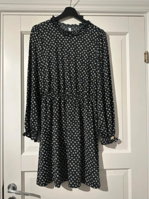 Marinblå kortklänning från Chiara Forti - Marinblå kortklänning från Chiara Forti med vitt prickigt mönster, volang vid halsringningen och långa ärmar med elastiska muddar. Klänningen har en avslappnad passform och är tillverkad i ett mjukt syntetmaterial. 