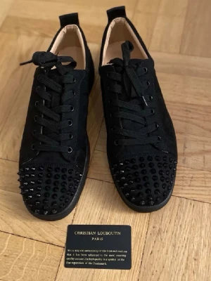 Christian Louboutin svarta sneakers med nitar - Svarta sneakers från Christian Louboutin med ikonisk röd sula och svarta nitar på tån. Skorna har snörning och är tillverkade i mocka med platt sula och rund tå. Snygg design med lyxig vibe och detaljer som sticker ut. Kommer med originalkartong och certifikat. Använda ett par gånger 
