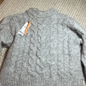 Ljusgrå kabelstickad tröja - Säljer en ljusgrå kabelstickad tröja med klassiskt flätmönster och ribbade muddar. Perfekt för kyliga dagar och riktigt mysig att bära. Passar dig som gillar en enkel men ändå snygg look.