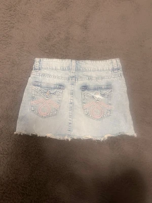 Ljusblå jeanskjol med broderade fickor - Säljer en ljusblå jeanskjol med rå kant och slitningar. Kjolen har två bakfickor med rosa broderade detaljer och knappstängning. Klassisk femficksmodell med bälteshällor och en kort passform som ger en cool vibe. Köpt från Gina tricot young stl 164.