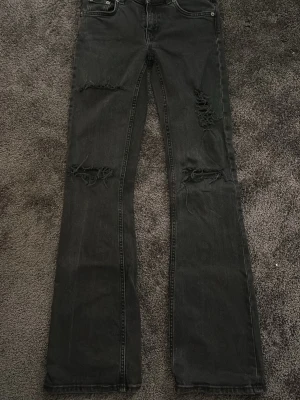 Svarta bootcut jeans från 157 - Bootcut jeans i svart denim från 157 med låg midja. Byxorna har slitna detaljer och hål på benen samt fransade benslut för en edgy vibe. Klassisk femficksmodell och dragkedja i gylfen. Perfekt för dig som gillar en avslappnad och trendig stil.