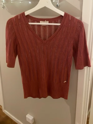 Mörkrosa ribbad topp med v-ringning - Säljer en snygg roströd topp med v-ringning och korta ärmar. Toppen har ett ribbat mönster och är stickad med tunna, genombrutna ränder som ger en luftig känsla. Perfekt till jeans eller kjol för en trendig look.