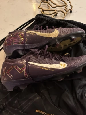 Nike Mercurial Air Zoom KM fotbollsskor - Säljer ett par Nike Mercurial Air Zoom KM fotbollsskor i mörklila med guldiga och vita detaljer. Skorna har snörning, platt sula med dobbar och coolt KM-mönster i lila och guld. Materialet är syntet och mesh för grym komfort och grepp på planen. Är i bra skick 