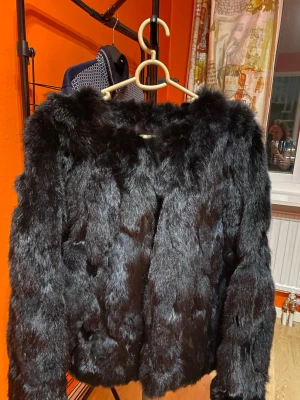 Svart pälsjacka från Skhoop XL - Säljer en svart pälsjacka från Skhoop i storlek XL. Jackan har en fluffig och mjuk känsla med långärmad design och klassisk rak passform. Perfekt för dig som vill ha en statementjacka med lyxig look och skön värme under kalla dagar.