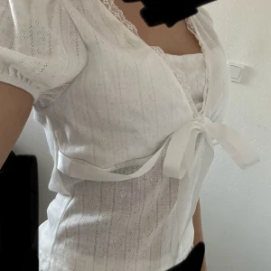 Vit omlottblus med rosett och spets - Säljer en söt vit omlottblus med korta puffärmar, spetskant vid v-ringningen och ett brett satinband som knyts framtill. Tror det är M men känns 100% som S & XS | VÄLJ GÄRNA INSTABOX | 💓