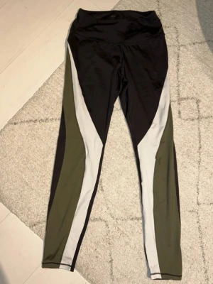 Svarta och gröna leggings från H&M - Snygga leggings från H&M med svart bas och coola gröna och vita paneler längs benen. Modellen är högmidjad och har en tight passform som sitter skönt mot kroppen. Perfekta för träning eller chill dagar. Materialet är stretchigt syntet för maximal komfort.