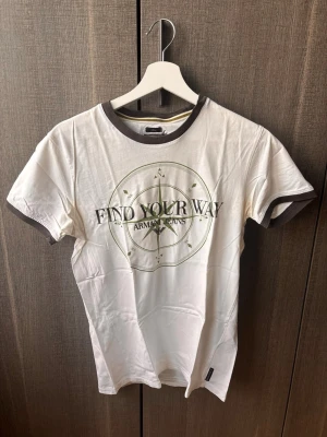 Vit t-shirt Armani Jeans med tryck - Vit t-shirt från Armani Jeans med grönt och svart kompass-tryck och texten 'Find Your Way'. Slim fit-modell med mörkgrå ribbad krage och ärmslut. Materialet är mjuk bomull och passformen är slim. Perfekt för dig som gillar stilrena detaljer och snygga prints. 