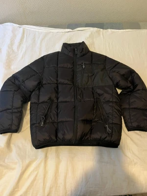 Svart pufferjacka med dragkedjor - Svart pufferjacka med quiltad design och hög krage. Jackan har tre praktiska fickor med dragkedja framtill och en stilren look. Materialet är syntet och jackan är perfekt för kyliga dagar. Storleken är M men den är liks liten som S.