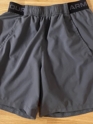 Grå träningsshorts från Under Armour - Säljer ett par grå träningsshorts från Under Armour med elastisk midja och diskret logga på linningen. Shortsen har en loose passform och är gjorda i lätt syntetmaterial som andas, perfekta för gymmet eller löprundan.