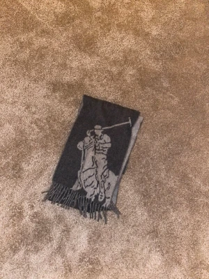 Grå och svart ullhalsduk Polo Ralph Lauren - Snygg halsduk från Polo Ralph Lauren i mjuk ull, med ett stort motiv av en polospelare. Färgen är mörkgrå och ljusgrå, och halsduken har klassiska fransar i ändarna. Perfekt för att addera lite stil till din vinteroutfit.