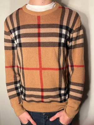 Beige rutig tröja från Burberry - Säljer en snygg beige tröja från Burberry med klassiskt rutmönster i svart, vitt och rött. Perfekt för dig som gillar stilrena och ikoniska plagg med lyxig känsla. Passar också storlek M     Grund värde 11 000kr
