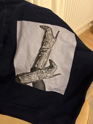 Mörkblå hoodie med glittrigt print - Säljer en mörkblå hoodie i storlek M med ett stort print på ryggen som föreställer glittriga stövlar och texten 'By Olivia'. Hoodien har snörning vid huvan och en klassisk magficka. Perfekt för dig som gillar coola detaljer. Märket är by Olivia som nu hittar på insta. Jag köpte den runt 800kr om jag minns rätt.