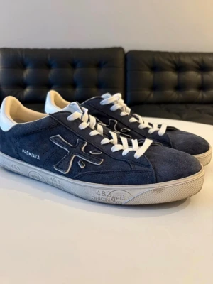 Premiata blå sneakers - Snygga sneakers från Premiata i mörkblå mocka med vita detaljer och snörning. Skorna har en rund tå, vit platt sula med grafiska tryck och en vit häl med Premiata-logga. Insidan är ljusbeige och yttermaterialet är mjuk mocka. Perfekt för dig som gillar stilrena och coola sneakers. Använd endast tre gånger. Priset kan diskuteras vid snabb affär.