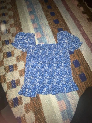 Blommig blå blus med volang - Supersöt blå blus med vitt blommönster och volangkant. Puffiga korta ärmar och smockad kropp ger en riktigt gullig och trendig look. Perfekt till jeans eller kjol för en fräsch sommarstil.