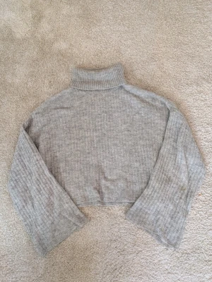 Grå croppad polotröja i stickat - Säljer en ljusgrå croppad polotröja i stickat material, med ribbad struktur och långa ärmar. Perfekt för dig som gillar en chill och trendig look med hög krage. Passar dig som vill ha något bekvämt och snyggt till hösten eller vintern. Ett hål i ena ärmen men inte så märkbart!