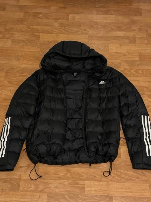 Svart pufferjacka från Adidas - Svart pufferjacka från Adidas med huva och klassiska vita ränder på ärmarna. Jackan har snörning i nederkant och dragkedja framtill. Perfekt för kyliga dagar och har en sportig vibe med Adidas-loggan på bröstet.