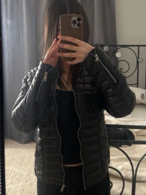 Svart quiltad pufferjacka med dragkedja - Säljer en svart pufferjacka med quiltad design och dragkedjor både framtill och vid ärmsluten. Jackan har en hög krage och en snygg, lite kortare passform som ger en modern vibe. Perfekt för dig som vill ha en stilren och varm jacka till höst och vinter.