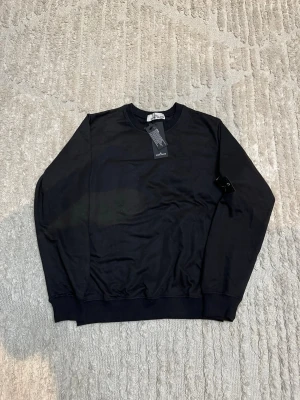 Svart sweatshirt från Stone Island - Svart sweatshirt från Stone Island med klassisk rund hals och ribbade muddar. Tröjan har den ikoniska Stone Island-loggan på vänster ärm och är tillverkad i mjuk bomull. Perfekt för dig som gillar stilrena och snygga plagg med streetkänsla.