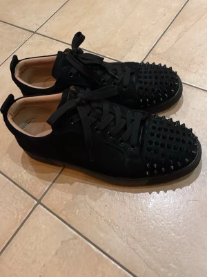 Svarta sneakers med nitar från Louboutin - Säljer ett par svarta sneakers från Louboutin med coola svarta nitar på tån och klassisk röd sula. Skorna har snörning och är gjorda i mocka med skinnfoder. Perfekt för dig som vill sticka ut med en edgy stil.