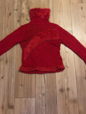 Röd polotröja med fluffiga detaljer - Säljer en röd polotröja med lång ärm och hög, fluffig krage. Tröjan har stickade och lurviga detaljer i samma färg över bröstet och längs nederkanten, vilket ger en unik look. Perfekt för dig som vill sticka ut lite extra.