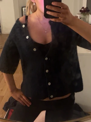 Mörkblå stickad kofta med knappar - Säljer en mörkblå stickad kofta med kortare ärmar och vita knappar framtill. Koftan har en loose fit och är perfekt att slänga över ett linne. Materialet är mjukt och mysigt, och modellen är lite boxig. Passar dig som gillar en chill och enkel stil.